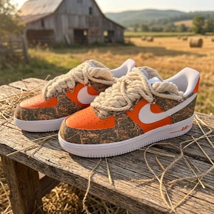 May include: A pair of low-top sneakers featuring bright orange leather toe boxes and side panels, accented by a brown and tan textured camouflage-like pattern. The shoes include thick, cream-colored braided rope laces and white Nike swoosh logos.