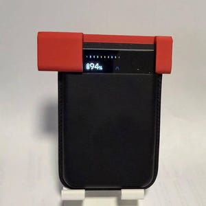 Puede incluir: Un cargador portátil rectangular de color negro con una funda protectora de silicona roja en el borde superior. El dispositivo cuenta con una pantalla digital que muestra un icono de batería, el texto 94% y barras indicadoras verticales.