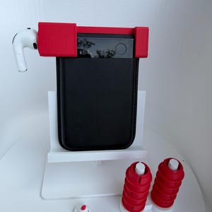 May include: A black rectangular power bank with a digital display showing 84% battery, held by a red silicone mount. A white wireless earbud is attached to the side. Two red cylindrical cable organizers and a small white chicken figurine are present.