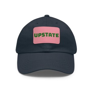 Könnte beinhalten: Eine marineblaue Baseballkappe mit einem rechteckigen, rosafarbenen Aufnäher in der Mitte des Vorderteils. Auf dem Aufnäher steht das Wort UPSTATE in fetten, dunkelgrünen Großbuchstaben. Die Kappe hat einen gebogenen Schirm und Ösen.