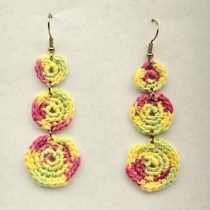 May include: A pair of drop earrings featuring three stacked, circular crochet motifs in variegated shades of yellow, lime green, and magenta. Each earring hangs from a thin, curved gold-tone metal hook, creating a vertical, layered design.
