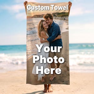 May include: A rectangular beach towel featuring a printed photograph of two people embracing on a sandy beach at sunset. The text Custom Towel is printed at the top, and the text Your Photo Here is printed in the center over the image.