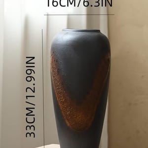 Może przedstawiać: Wysoki, cylindryczny wazon z matowym czarnym wykończeniem i teksturowanym, rdzawym akcentem w kształcie litery V. Wazon ma 16 cm szerokości i 33 cm wysokości, posiada gładką górną krawędź oraz zwężającą się podstawę.