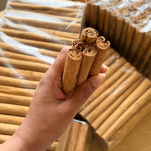 May include: A collection of light brown cinnamon sticks with a tubular, rolled bark texture. Several individual sticks are held together in a cluster, while additional bundles of cinnamon sticks are packaged in clear plastic wrap in the background.