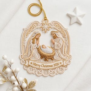 May include: An oval-shaped cream and gold embroidered Christmas ornament featuring a nativity scene with figures kneeling around a manger. A banner at the bottom displays the text Merry Christmas 2026. A gold cord is attached for hanging.