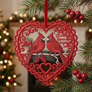 Pode incluir: Um ornamento bordado em forma de coração vermelho com dois cardeais pousados num ramo com bagas. O design inclui bordas intrincadas semelhantes a renda e o texto "Cardinals appear when angels are near" bordado em fio vermelho sobre tecido.