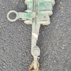 May include: An antique green and silver metal clamp with a rope attached. The clamp has a ring on one side and a lever on the other. The rope is brown and knotted.