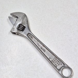 May include: A silver-colored metal adjustable wrench with a knurled adjustment screw. The handle is stamped with the text 6 IN., 706 PROTO, and FORGED ALLOY STEEL. The tool features a movable jaw and a circular hole at the end of the handle.