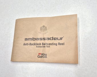 Puede incluir: Un manual de instrucciones beige para un carrete de baitcasting anti-retroceso Ambassadeur. La portada presenta el nombre de la marca, la descripción del producto y el logotipo de Abu Garcia. Un escudo está impreso en la parte superior.