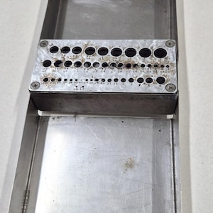 May include: A stainless steel drill bit index with multiple sized holes, arranged in rows. The index is set inside a rectangular tray. The drill bit sizes are marked on the index. The tray and index show signs of wear.