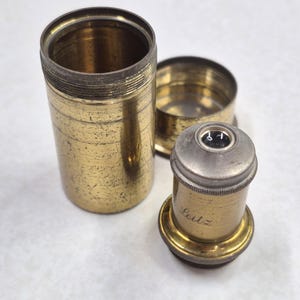 May include: A vintage brass microscope objective with a silver top and the engraved word "Leitz". The objective is next to its brass storage case, which is in three pieces. The case has a cylindrical shape.