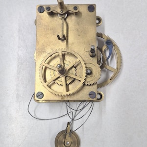 Puede incluir: Un movimiento de reloj antiguo con engranajes de lat&oacute;n y un peso de p&eacute;ndulo. El mecanismo rectangular muestra el texto "CHELSEA CLOCK CO. BOSTON, U.S.A." y "60389". Los intrincados engranajes resaltan la mec&aacute;nica interna del reloj.