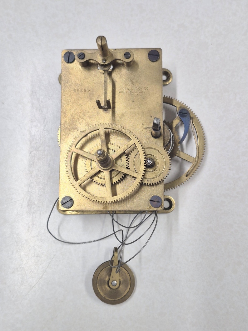 Puede incluir: Un movimiento de reloj antiguo con engranajes de lat&oacute;n y un peso de p&eacute;ndulo. El mecanismo rectangular muestra el texto "CHELSEA CLOCK CO. BOSTON, U.S.A." y "60389". Los intrincados engranajes resaltan la mec&aacute;nica interna del reloj.