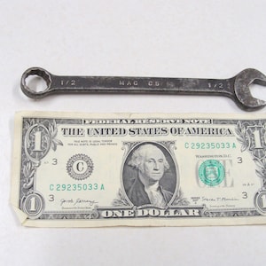 May include: A vintage metal wrench with the markings "MAC C5" and "1/2" is displayed next to a one-dollar bill. The wrench is a dark gray color, and the bill features a portrait of George Washington.