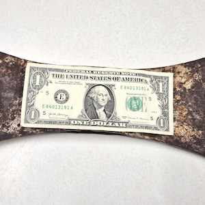 May include: A double-bit axe head with a heavily rusted, dark brown and metallic finish. A United States one-dollar bill is placed horizontally across the center of the axe head to provide a visual scale reference for the object's size.