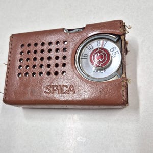 May include: A vintage brown leather-cased radio with the brand name "SPICA" printed on the front. The radio features a circular dial with numbers and a red and silver center. The case has a series of small holes and shows signs of wear.