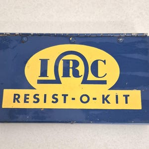 May include: A rectangular blue metal storage box with a hinged lid. The front features a yellow oval containing the dark blue letters IRC and a yellow rectangular banner below with the dark blue text RESIST-O-KIT printed in a sans-serif font.