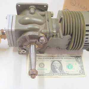 May include: An olive-drab metal mechanical component with a cylindrical cooling element and a protruding shaft. A one-dollar bill is placed beneath the component for scale. A yellow tag is attached to a brass fitting.