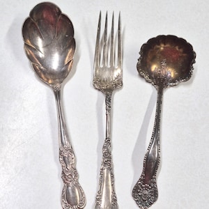 May include: A set of three silver-toned utensils consisting of a large scalloped serving spoon, a four-tined dinner fork, and a smaller scalloped bowl spoon. Each piece features intricate embossed floral and scrollwork detailing on the handles.