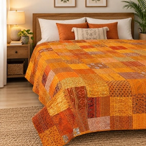 Könnte beinhalten: Eine Patchwork-Tagesdecke aus verschiedenen orangefarbenen, gelben und braunen Stoffquadraten mit unterschiedlichen Blumen- und Geometriemustern. Die Decke ist mit sichtbaren weißen Fäden abgesteppt und liegt auf einem Bett mit weißen und orangefarbenen Kissen.