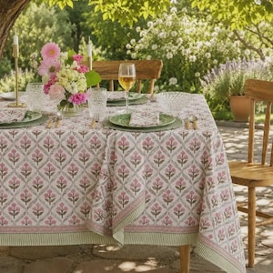 May include: A rectangular tablecloth featuring a repeating diamond pattern with pink floral motifs and green stems. The fabric has a striped border in shades of green and white. The table is set with clear glassware, plates, and a floral centerpiece.