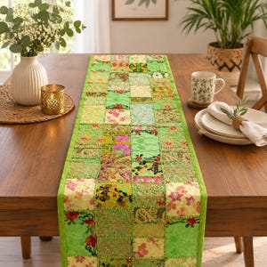 Op de afbeelding: Een rechthoekige tafelloper met een patchworkontwerp van diverse groene, gele en roze stoffen vierkanten. De loper is samengesteld uit meerdere getextureerde stoffen met ingewikkelde bloemen- en paisleypatronen, metalen borduursels en lovertjes.