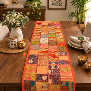Op de afbeelding: Een rechthoekige tafelloper met een patchworkontwerp van diverse stoffen vierkanten in levendige kleuren zoals oranje, roze, blauw en goud. De loper is versierd met ingewikkeld borduurwerk, kleine kraaltjes en stoffen bloemapplicaties.