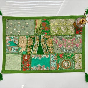 May include: A rectangular green fabric mat featuring a patchwork design of various embroidered textiles in shades of lime, emerald, and olive green. The mat is accented with sequins, gold thread, and small tassels at each corner, with a glass diffuser.