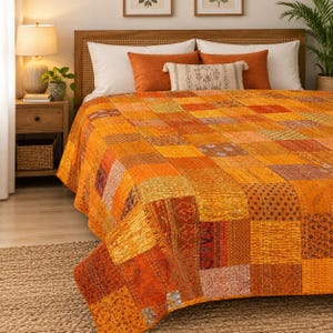 Op de afbeelding: Een patchwork sprei bestaande uit diverse oranje, gele en bruine stoffen vierkanten met uiteenlopende bloem- en geometrische patronen. De sprei is voorzien van zichtbaar wit stiksel en ligt op een bed met witte en oranje kussens.