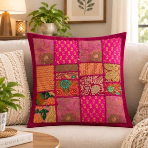 May include: A square throw pillow featuring a vibrant patchwork design with various shades of pink, magenta, and gold. The fabric panels display intricate embroidered floral patterns, metallic thread accents, and textured fabric squares.