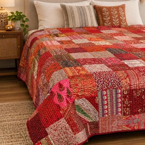 May include: A patchwork quilt featuring a vibrant array of red, pink, and orange fabric squares with intricate floral and geometric patterns. The quilt is constructed with visible running stitch quilting details throughout the entire surface area.