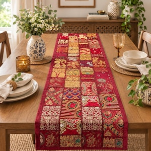 May include: A rectangular table runner featuring a patchwork design of red fabric squares. Each square is decorated with intricate gold embroidery, metallic thread work, and small reflective mirror accents, creating a vibrant, textured surface.