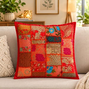 Op de afbeelding: Een vierkant sierkussen met een levendig patchworkontwerp van rode, oranje en roze stoffen vierkanten. Het oppervlak is versierd met ingewikkeld goudborduurwerk, bloemmotieven en gestructureerde stiksels, omlijst door een effen rode rand.