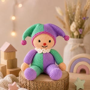 May include: A knitted doll featuring a jester-style hat and a two-toned outfit in alternating purple and mint green. The doll has a peach-coloured face, a small red nose, a stitched smile, and a white ruffled collar around its neck.