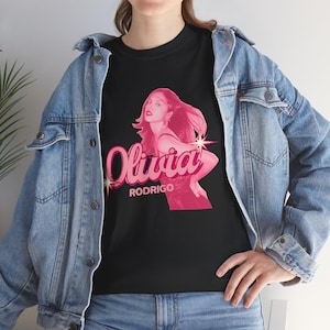May include: A black short-sleeved t-shirt featuring a pink graphic print of Olivia Rodrigo. The design includes the name Olivia in a stylized script font with star accents, the word RODRIGO in block letters, and a portrait of the artist.