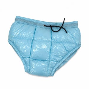 Op de afbeelding: Een lichtblauwe gewatteerde slip in donsjack-stijl met een glanzende, reflecterende afwerking. Het kledingstuk heeft een brede, elastische tailleband met een dun zwart trekkoord dat in het midden aan de voorzijde tot een lusje is gestrikt.