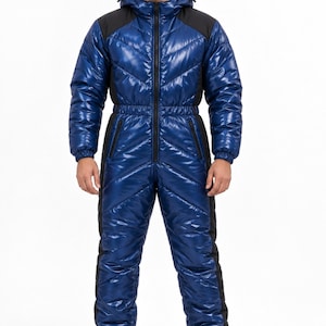 Op de afbeelding: Een glanzende blauwe gewatteerde jumpsuit met een capuchon, een ritssluiting over de hele lengte aan de voorzijde en zwarte zijpanelen. Het kledingstuk heeft lange mouwen met elastische boorden, een getailleerde taille en lange broekspijpen met elastische enkels, gecombineerd met zwarte sneakers.