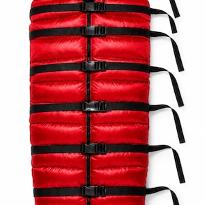 May include: A bright red, quilted, hooded sleeping bag featuring a vertical central zip and seven horizontal black nylon straps with plastic side-release buckles. The interior of the hood is lined with a shiny, smooth black material.