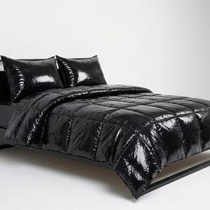 Puede incluir: Una estructura de cama negra con un edredón acolchado negro a juego de alto brillo y dos almohadas rectangulares. La ropa de cama presenta un acabado brillante y reflectante con un patrón de costuras en cuadrícula, creando un aspecto inflado.