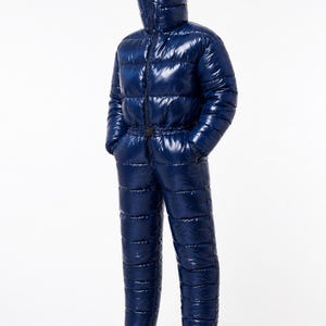 Puede incluir: Un mono acolchado de cuerpo entero en color azul marino brillante, con una capucha de cuello alto que se cierra hasta cubrir el rostro, mangas largas y puños elásticos en los tobillos. La prenda presenta un diseño acolchado horizontal y un cinturón negro.