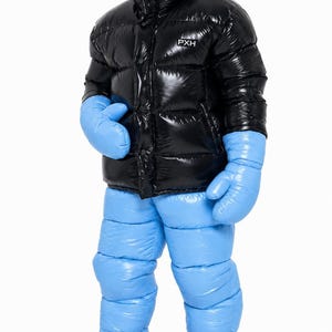 Op de afbeelding: Een glanzend zwart donsjack met een hoge kraag en de tekst PXH op de borst, gecombineerd met een lichtblauwe donsbroek, bijpassende lichtblauwe oversized wanten en felroze lage schoenen met zwarte zolen en veters.