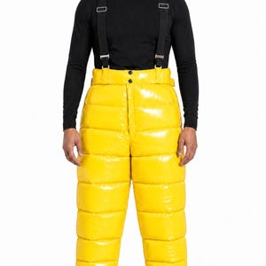 Puede incluir: Pantalones acolchados de color amarillo brillante de talle alto con acabado brillante y costuras horizontales. Los pantalones cuentan con tirantes negros integrados con herrajes metálicos plateados, cintura abotonada y puños elásticos en los tobillos.