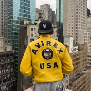 Può includere: Una giacca giallo brillante con scritte nere che recitano AVIREX USA, caratterizzata da una toppa circolare con la bandiera americana e il testo ALL AMERICANS ESTABLISHED 1975. La giacca è abbinata a un berretto da baseball blu scuro con logo NY bianco.