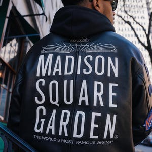 May include: A black leather jacket featuring a white graphic on the back that reads MADISON SQUARE GARDEN, THE WORLD'S MOST FAMOUS ARENA, with an architectural line drawing above the text and the brand name AVIREX printed on the hood.