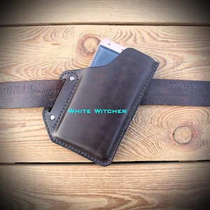 Puede incluir: Una funda de cuero marrón oscuro para teléfono con costuras visibles y dos remaches metálicos, insertada en un cinturón de cuero marrón oscuro. El texto WHITE WITCHER está impreso en azul claro en la parte frontal de la funda, que contiene un smartphone.