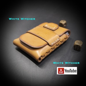 Può includere: Un portafoglio in pelle color cuoio con chiusura a patta e cuciture a vista, accompagnato da due dadi metallici. Il testo "WHITE WITCHER" appare due volte in ciano e un'icona "PDF YouTube" è visibile nell'angolo dell'immagine.