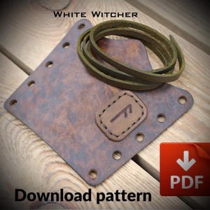 Può includere: Un pezzo di pelle marrone quadrato con texture maculata, fori lungo i bordi e una piccola toppa rettangolare con un simbolo runico. Un cordino in pelle verde oliva arrotolato è appoggiato sopra. Testo: WHITE WITCHER e PDF.