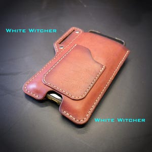 Può includere: Un porta carte in pelle marrone con cuciture a vista e una tasca frontale. Il testo "WHITE WITCHER" è stampato in azzurro nell'angolo in alto a sinistra e in basso a destra. Il design include un punto di fissaggio in metallo nella parte superiore.