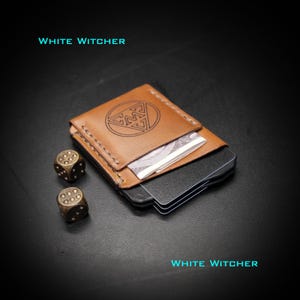 May include: A tan leather card holder with visible stitching and a circular logo featuring the letters MW. The wallet contains several black cards and is accompanied by two metallic bronze-colored dice. The text WHITE WITCHER appears twice.