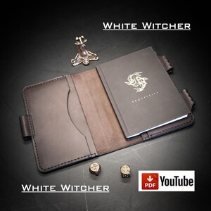 Könnte beinhalten: Ein dunkelbrauner Notizbuchumschlag aus Leder mit genähten Kanten, der ein dunkelgraues Notizbuch mit goldenem Koi-Fisch-Emblem und dem Text PROSPERITY enthält. Beinhaltet eine kleine Froschfigur aus Metall, zwei Würfel und den Text WHITE WITCHER.